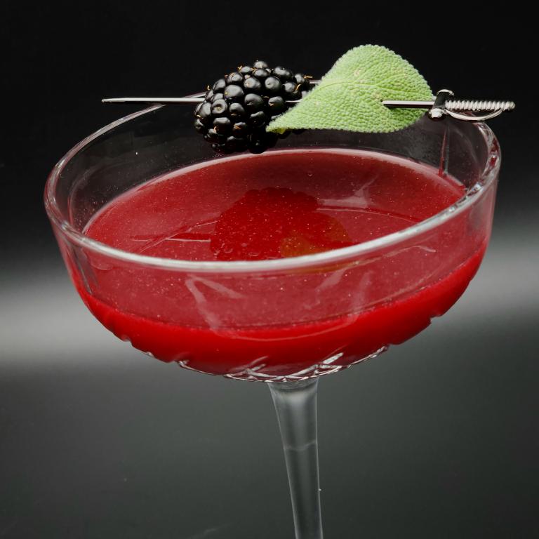 large coupe half-filled with dark red liquid. Garnished with a small metal sword skewering a sage leaf and a blackberry.